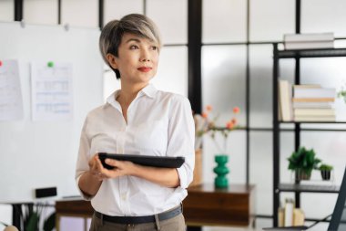 Bir kadın mimar, tasarım kitapları ve materyallerle dolu modern bir ofiste fikirlerini sunmak için özenle bir tablet tutuyor..
