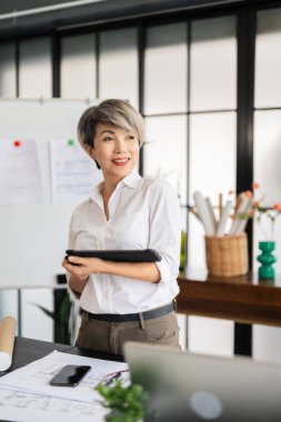 Bir kadın mimar, tasarım kitapları ve materyallerle dolu modern bir ofiste fikirlerini sunmak için özenle bir tablet tutuyor..
