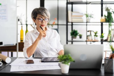 Bir kadın mimar masasında oturmuş dizüstü bilgisayarındaki tasarım planlarını inceliyor. Tasarım malzemeleri ve proje belgeleriyle çevrili..