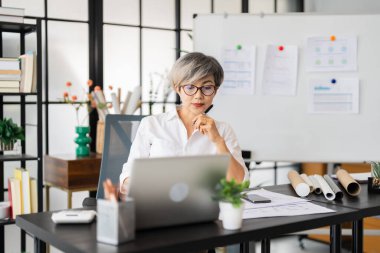 Bir kadın mimar masasında oturmuş dizüstü bilgisayarındaki tasarım planlarını inceliyor. Tasarım malzemeleri ve proje belgeleriyle çevrili..