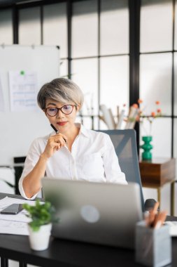 Bir kadın mimar masasında oturmuş dizüstü bilgisayarındaki tasarım planlarını inceliyor. Tasarım malzemeleri ve proje belgeleriyle çevrili..