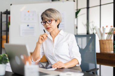 Bir kadın mimar masasında oturmuş dizüstü bilgisayarındaki tasarım planlarını inceliyor. Tasarım malzemeleri ve proje belgeleriyle çevrili..