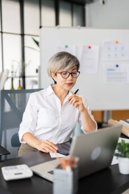 Bir kadın mimar masasında oturmuş dizüstü bilgisayarındaki tasarım planlarını inceliyor. Tasarım malzemeleri ve proje belgeleriyle çevrili..