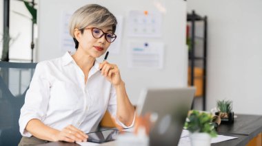 Bir kadın mimar masasında oturmuş dizüstü bilgisayarındaki tasarım planlarını inceliyor. Tasarım malzemeleri ve proje belgeleriyle çevrili..