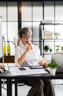 Dişi bir mimar, elinde kalemle masasının planlarını incelerken, iyi organize edilmiş bir çalışma alanına dizayn edilmiş materyallerle derin düşüncelere dalmıştır..