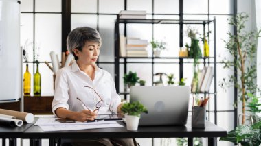Dişi bir mimar, elinde kalemle masasının planlarını incelerken, iyi organize edilmiş bir çalışma alanına dizayn edilmiş materyallerle derin düşüncelere dalmıştır..