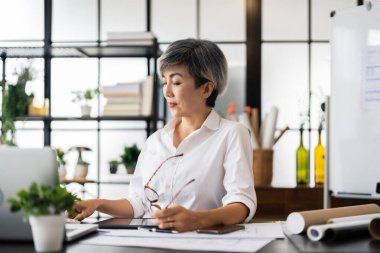 Dişi bir mimar, elinde kalemle masasının planlarını incelerken, iyi organize edilmiş bir çalışma alanına dizayn edilmiş materyallerle derin düşüncelere dalmıştır..