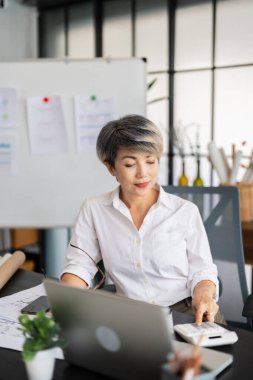 Modern ofisinde çeşitli mimari materyaller ve planlarla çevrili tasarım planları üzerinde çalışırken bir kadın mimar dizüstü bilgisayarına odaklanır.