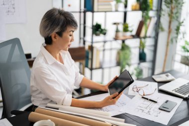 Bir kadın mimar, masasında otururken tabletinin mimari planlarını gözden geçirir. Etrafında çeşitli tasarım çizimleri ve araçlar vardır. Modern, iyi düzenlenmiş bir ofiste..