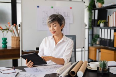 Bir kadın mimar, masasında otururken tabletinin mimari planlarını gözden geçirir. Etrafında çeşitli tasarım çizimleri ve araçlar vardır. Modern, iyi düzenlenmiş bir ofiste..