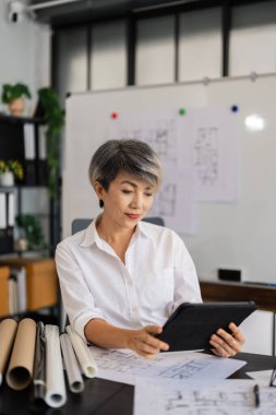 Bir kadın mimar, masasında otururken tabletinin mimari planlarını gözden geçirir. Etrafında çeşitli tasarım çizimleri ve araçlar vardır. Modern, iyi düzenlenmiş bir ofiste..