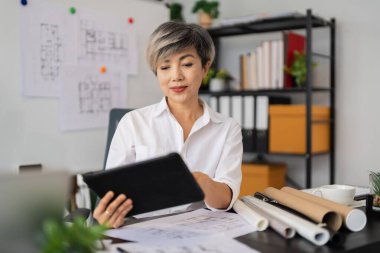 Bir kadın mimar, masasında otururken tabletinin mimari planlarını gözden geçirir. Etrafında çeşitli tasarım çizimleri ve araçlar vardır. Modern, iyi düzenlenmiş bir ofiste..