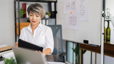 Bir kadın mimar, masasında otururken tabletinin mimari planlarını gözden geçirir. Etrafında çeşitli tasarım çizimleri ve araçlar vardır. Modern, iyi düzenlenmiş bir ofiste..