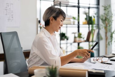 Bir kadın mimar, masasında otururken tabletinin mimari planlarını gözden geçirir. Etrafında çeşitli tasarım çizimleri ve araçlar vardır. Modern, iyi düzenlenmiş bir ofiste..