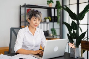 Dişi bir mimar dizüstü bilgisayarı üzerinde çalışır, planlar ve mimari çizimlerle çevrilidir, düzenli ve modern ofis ortamında tasarım projelerine yoğunlaşır..