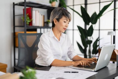 Dişi bir mimar dizüstü bilgisayarı üzerinde çalışır, planlar ve mimari çizimlerle çevrilidir, düzenli ve modern ofis ortamında tasarım projelerine yoğunlaşır..