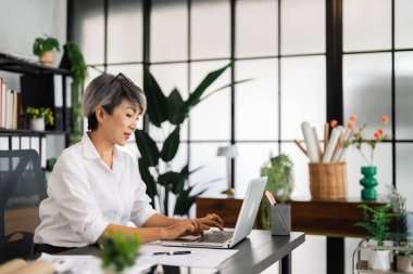 Dişi bir mimar dizüstü bilgisayarı üzerinde çalışır, planlar ve mimari çizimlerle çevrilidir, düzenli ve modern ofis ortamında tasarım projelerine yoğunlaşır..