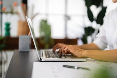Dişi bir mimar dizüstü bilgisayarı üzerinde çalışır, planlar ve mimari çizimlerle çevrilidir, düzenli ve modern ofis ortamında tasarım projelerine yoğunlaşır..
