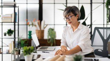 Beyaz gömlekli bir kadın mimar, modern, iyi organize edilmiş bir ofiste, araçlar ve planlar ile çevrili, planlar ve planlar üzerinde, planlar ve planlar üzerinde, çalışma masasında özenle çalışıyor.