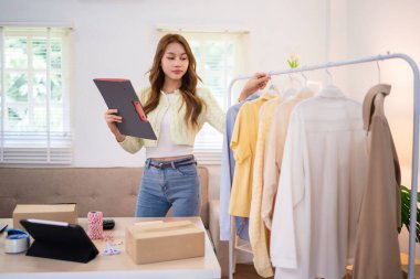 Çevrimiçi kıyafet işini yöneten genç bir kadın giyim mağazasının yanında dururken elinde bir tablet tutuyor. Siparişleri düzenliyor ya da kontrol ediyor gibi görünüyor., 