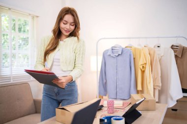 Online dükkanını işleten genç bir kadın tabletini kontrol ederken gülümsüyor. Elbise rafının yanında duruyor. Masasında kutular ve nakliye malzemeleri var.,