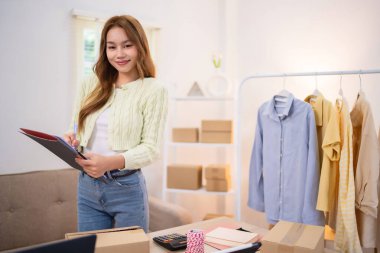 Online dükkanını işleten genç bir kadın tabletini kontrol ederken gülümsüyor. Elbise rafının yanında duruyor. Masasında kutular ve nakliye malzemeleri var.,