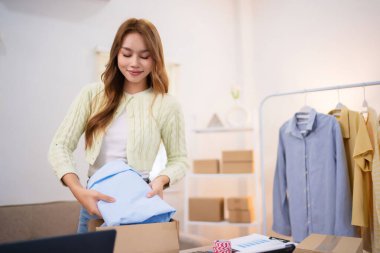 Çevrimiçi kıyafet tüccarı olarak çalışan genç bir kadın masasında paketler hazırlıyor. Etrafı dizüstü bilgisayarlar, kutular ve giysi parçalarıyla çevrili..