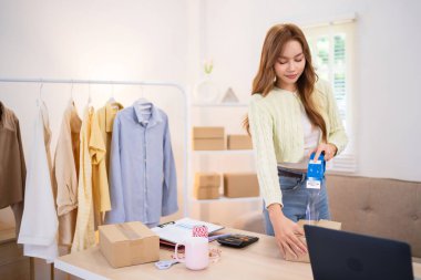 Genç bir kadın çevrimiçi giyim mağazası için sipariş hazırlarken bir kutuyu bant ile mühürlüyor. Etrafında nakliye malzemeleri ve dizüstü bilgisayarlarla aydınlık bir odada.