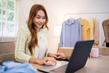 Çevrimiçi giyim sektörü işleten genç bir kadın masadaki bilgisayarını kullanırken gülümsüyor. Etrafı giysi parçalarıyla çevrili ve emirlerini uyguluyor. 