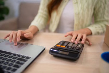 Çevrimiçi giyim sektörü işleten genç bir kadın masadaki bilgisayarını kullanırken gülümsüyor. Etrafı giysi parçalarıyla çevrili ve emirlerini uyguluyor. 