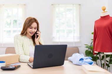 Genç bir kadın dizüstü bilgisayarı üzerinde çalışırken telefonda konuşuyor. İnternetteki kıyafet işini yönetiyor. Parlak bir odada.,