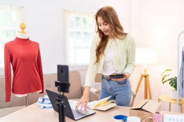 Genç bir kadın masasında duruyor, telefonunu kullanarak online giyim işi için çalışma alanını düzenliyor. Etrafında ambalaj malzemeleri ve kıyafetlerle içeriği çekiyor..
