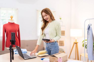 Genç bir kadın masasında duruyor, telefonunu kullanarak online giyim işi için çalışma alanını düzenliyor. Etrafında ambalaj malzemeleri ve kıyafetlerle içeriği çekiyor..