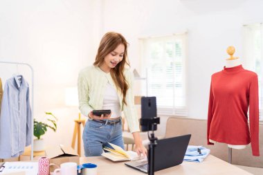 Genç bir kadın masasında duruyor, telefonunu kullanarak online giyim işi için çalışma alanını düzenliyor. Etrafında ambalaj malzemeleri ve kıyafetlerle içeriği çekiyor..