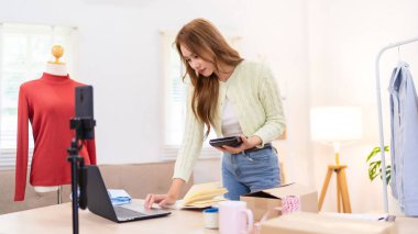 Genç bir kadın masasında duruyor, telefonunu kullanarak online giyim işi için çalışma alanını düzenliyor. Etrafında ambalaj malzemeleri ve kıyafetlerle içeriği çekiyor..