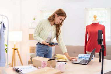 Genç bir kadın masasında duruyor, telefonunu kullanarak online giyim işi için çalışma alanını düzenliyor. Etrafında ambalaj malzemeleri ve kıyafetlerle içeriği çekiyor..