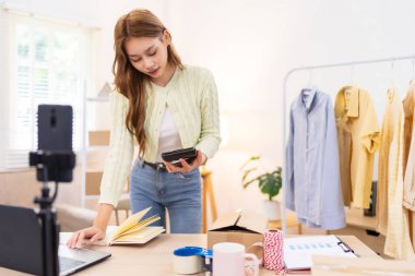 Genç bir kadın masasında duruyor, telefonunu kullanarak online giyim işi için çalışma alanını düzenliyor. Etrafında ambalaj malzemeleri ve kıyafetlerle içeriği çekiyor..