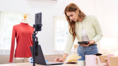 Genç bir kadın masasında duruyor, telefonunu kullanarak online giyim işi için çalışma alanını düzenliyor. Etrafında ambalaj malzemeleri ve kıyafetlerle içeriği çekiyor..