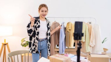 Genç bir kadın çalışma alanında elinde mavi bir tişörtle dikiliyor ve bunu online giyim işi için sergiliyor. İçeriği filme alıyor. 