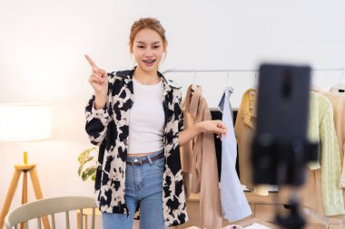 Genç bir kadın çalışma alanında elinde mavi bir tişörtle dikiliyor ve bunu online giyim işi için sergiliyor. İçeriği filme alıyor. 