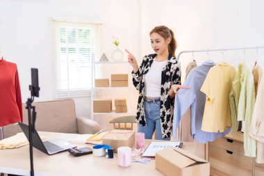 Genç bir kadın çalışma alanında elinde mavi bir tişörtle dikiliyor ve bunu online giyim işi için sergiliyor. İçeriği filme alıyor. 