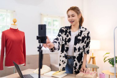 Genç bir kadın çevrimiçi giyim işi için içeriğini filme almaya hazırlanırken kamerasını ayarlıyor. O, arka planda kırmızı bir kazak gösteren bir mankenle gülümsüyor..