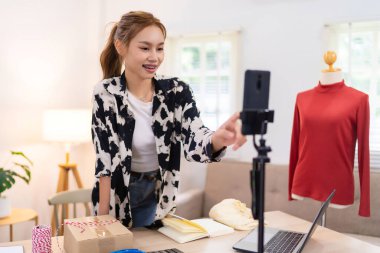 Genç bir kadın çevrimiçi giyim işi için içeriğini filme almaya hazırlanırken kamerasını ayarlıyor. O, arka planda kırmızı bir kazak gösteren bir mankenle gülümsüyor..