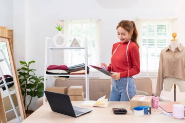 Genç bir kadın elinde bir tablet tutuyor ve çevrimiçi giyim işi için içeriği çekmeye hazırlanıyor. Kırmızı bir kazak giyiyor ve düzenli bir çalışma alanında çalışıyor.