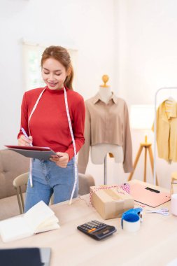 Genç bir kadın elinde bir tablet tutuyor ve çevrimiçi giyim işi için içeriği çekmeye hazırlanıyor. Kırmızı bir kazak giyiyor ve düzenli bir çalışma alanında çalışıyor.