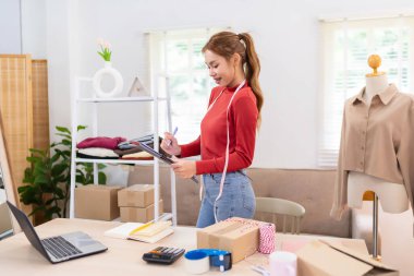 Genç bir kadın elinde bir tablet tutuyor ve çevrimiçi giyim işi için içeriği çekmeye hazırlanıyor. Kırmızı bir kazak giyiyor ve düzenli bir çalışma alanında çalışıyor.