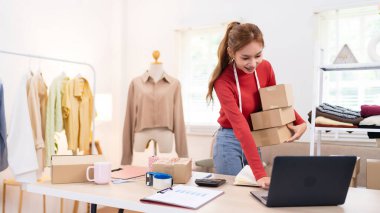Çevrimiçi giyim işi için siparişleri hazırlayan genç bir kadın elinde birkaç kutu tutuyor. Gülümsüyor ve düzenli bir çalışma alanında çalışıyor.
