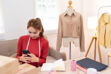 Genç bir kadın masasında oturuyor ve internetteki kıyafet işlerinin siparişlerini yönetmek için telefonunu kullanıyor. Etrafı kutular, defterler ve paketleme malzemeleriyle çevrili.
