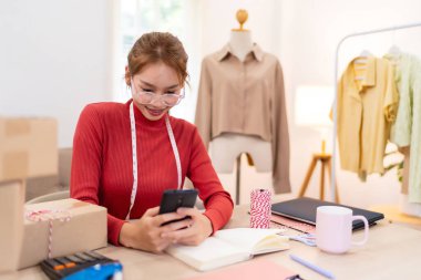 Genç bir kadın masasında oturuyor ve internetteki kıyafet işlerinin siparişlerini yönetmek için telefonunu kullanıyor. Etrafı kutular, defterler ve paketleme malzemeleriyle çevrili.