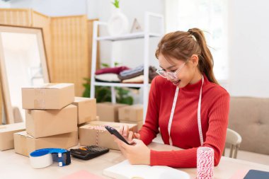 Genç bir kadın masasında oturuyor ve internetteki kıyafet işlerinin siparişlerini yönetmek için telefonunu kullanıyor. Etrafı kutular, defterler ve paketleme malzemeleriyle çevrili.